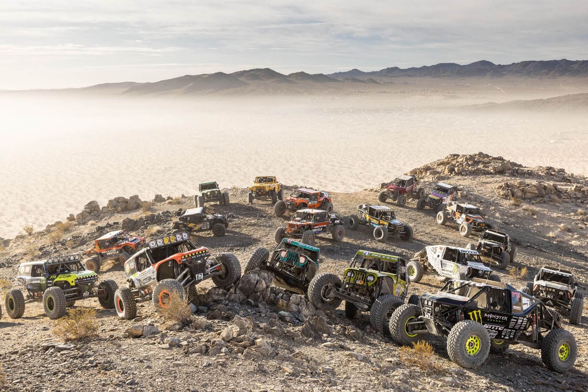 Group of rock crawler off-road vehicles lined up on a rocky hill during King of the Hammers race,
with a desert landscape and mountains in the background.